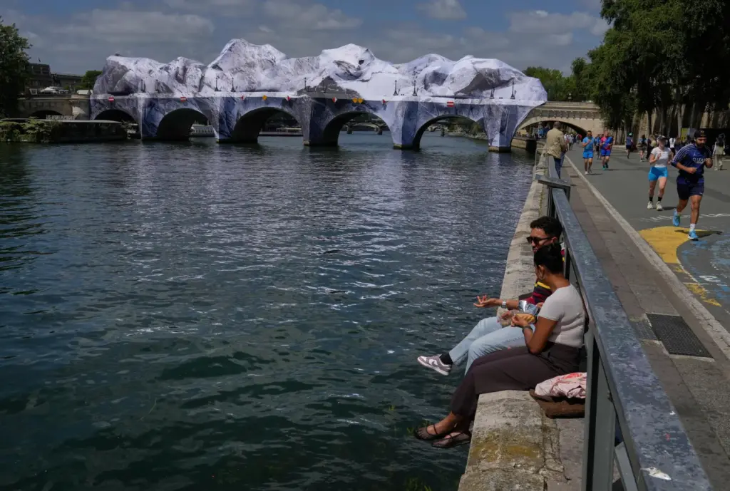 People sit along the Seine river next to “The Pont...