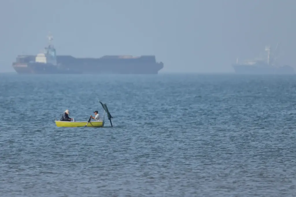 Two men sit in a small boat as cargo ships are anchored in the Strait of Hormuz.