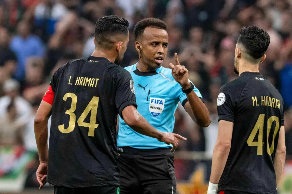 Referee Omar Artan is confronted by players after calling a penalty kick.
