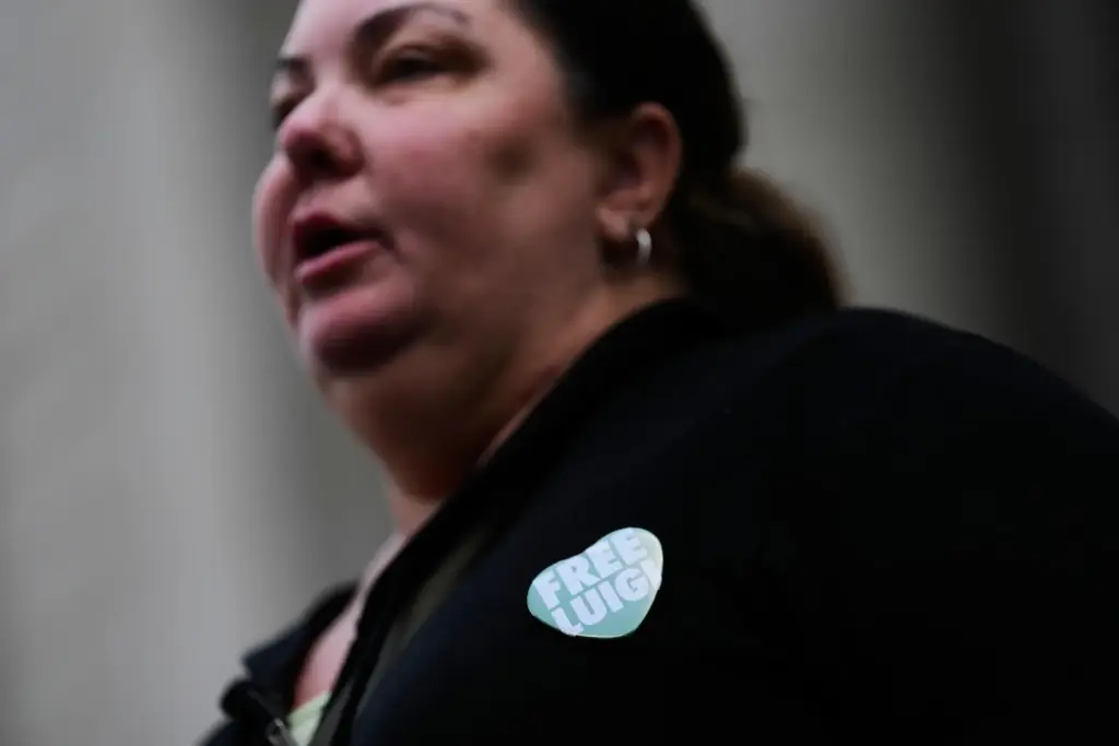 A woman wearing a “Free Luigi” pin talks to reporters...