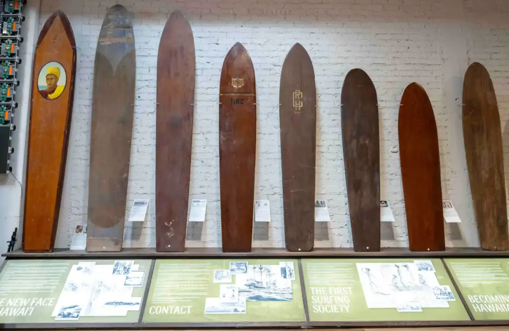 A collection of antique surfboards, mostly made of wood, hang...