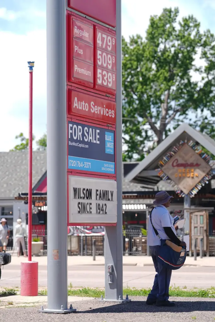 A United States Postal Service postman stands in the shade of a gas station sign.