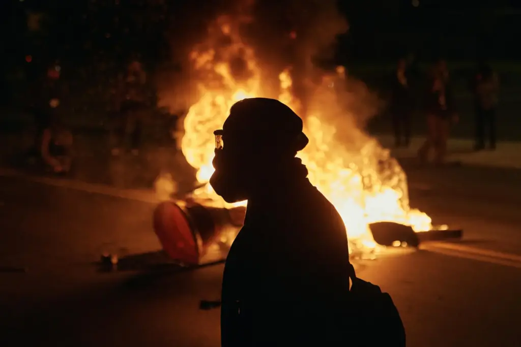 A man wearing protective gear walks in front of a burning barricade.