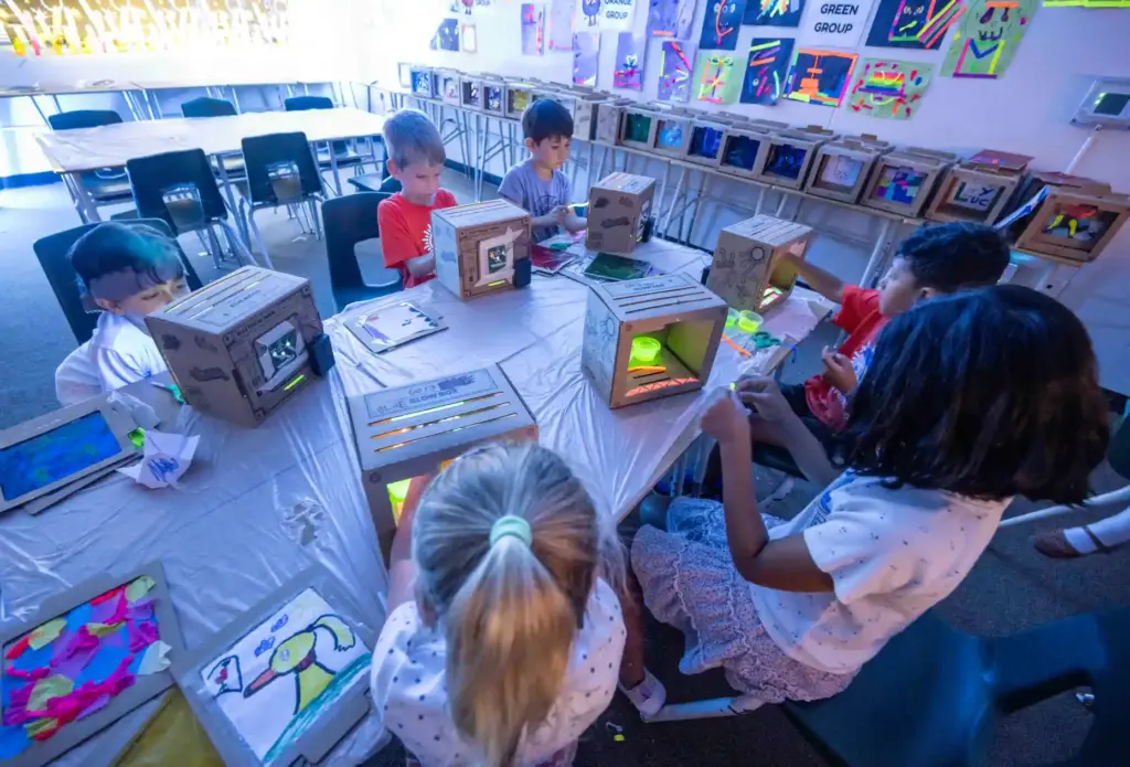 Camp Invention campers sit together in a blacklight-lit classroom at...