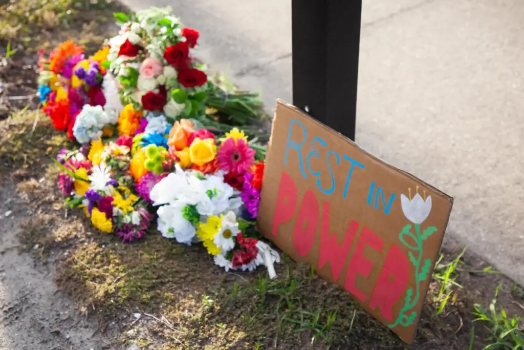 A sign reading “rest in power” is displayed alongside flowers...