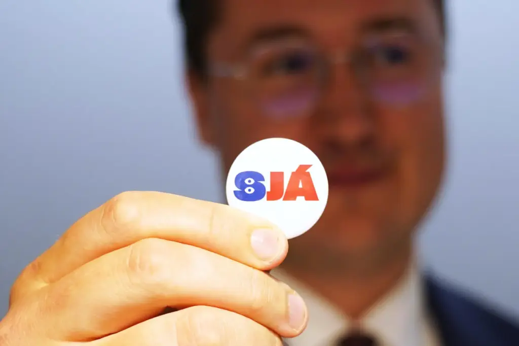 A man holds a badge reading “See”, at the opening...