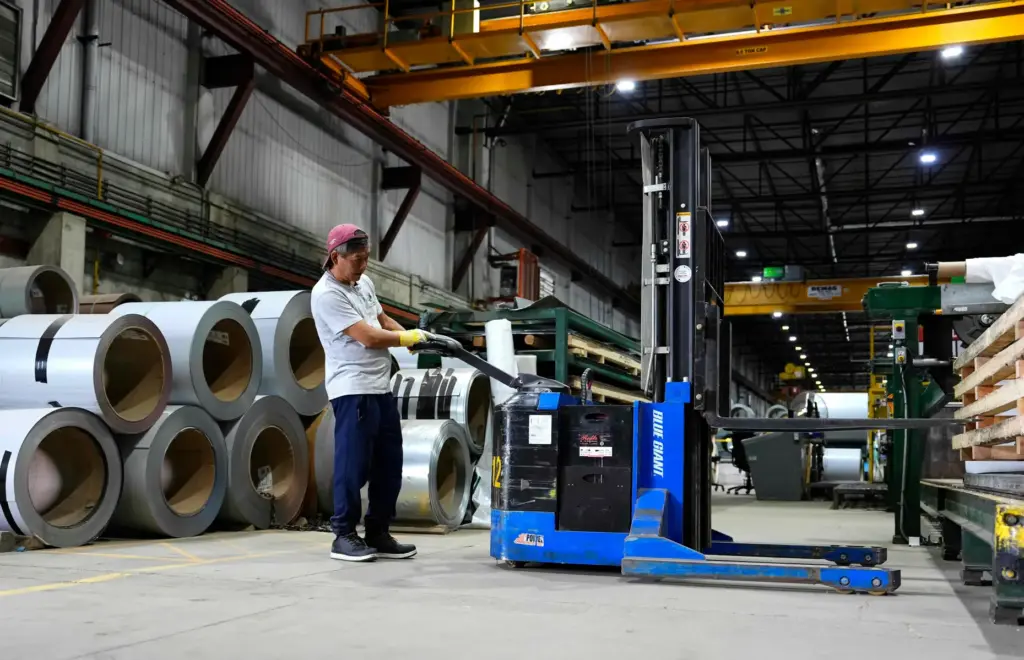 A worker operates a forklift in front of steel coils.