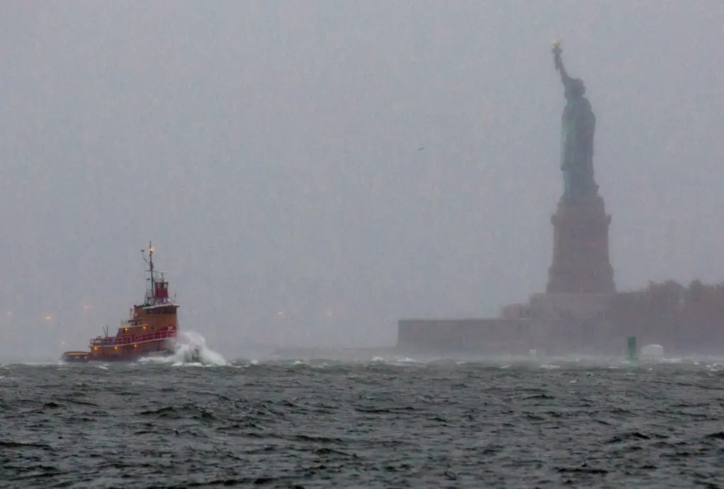 FILE – Waves crash over the bow of a tug...