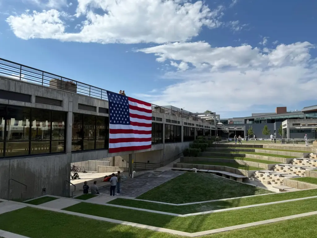 People gaze down at the site where Charlie Kirk was shot, above which hangs an American flag.