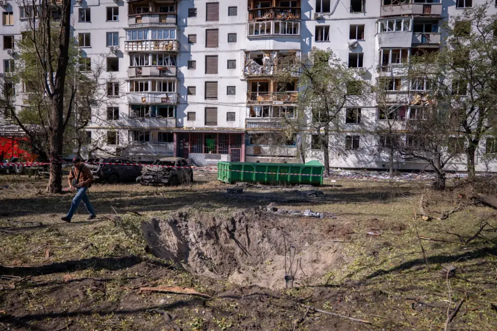 A missile crater is seen in the foreground of a damaged apartment building.