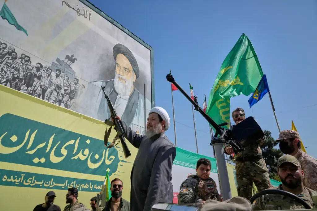 An armed Shiite cleric rides aboard a military vehicle past a billboard depicting Ayatollah Ruhollah Khomeini.
