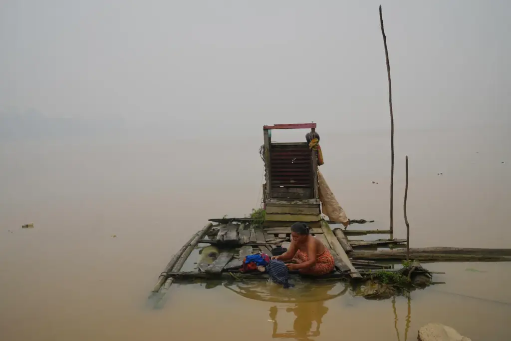 A woman washes clothes on a floating wooden platform along...