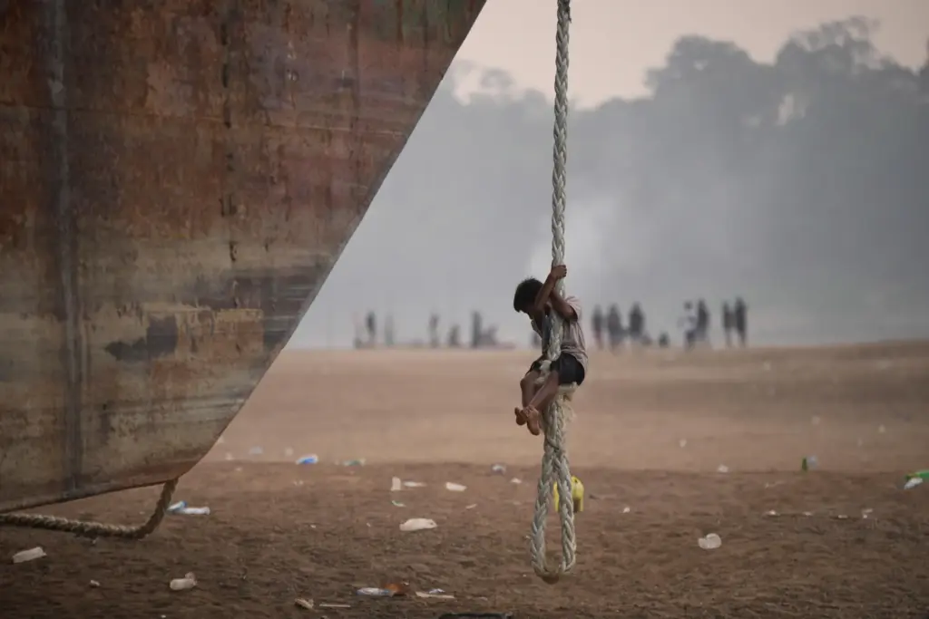 A boy climbs a rope hanging from a barge stranded...