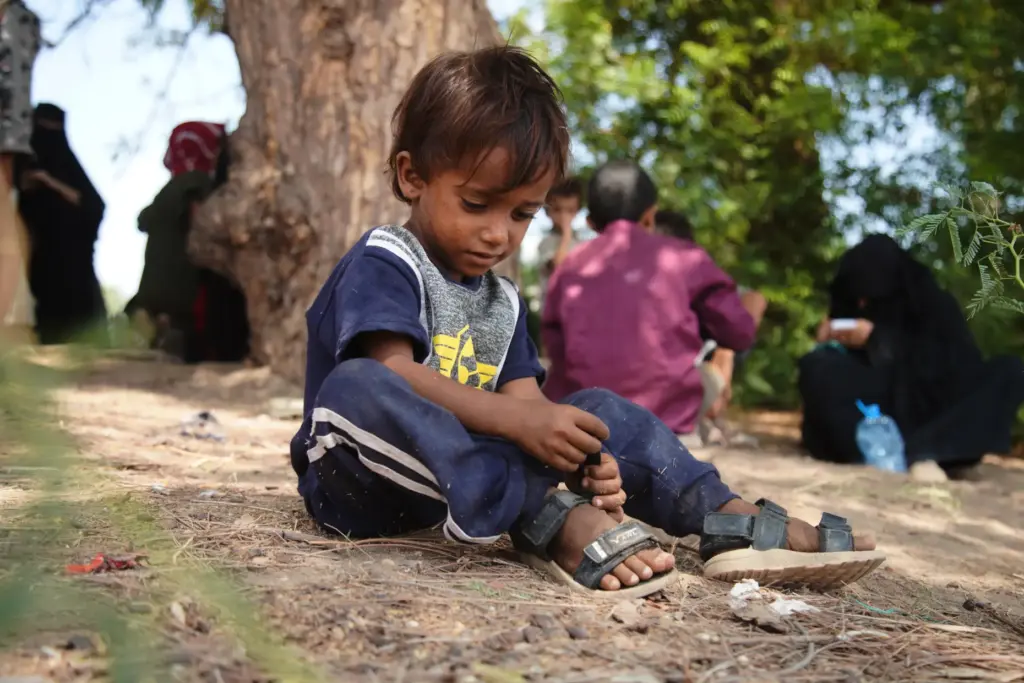 A displaced Yemeni child sits under a tree as families gather to receive aid.
